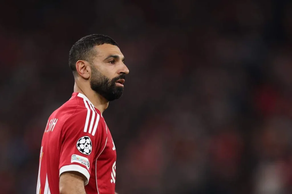 Side profile of a bearded male footballer in a red Liverpool jersey standing on the pitch during a match, with a blurred stadium crowd in the background.
