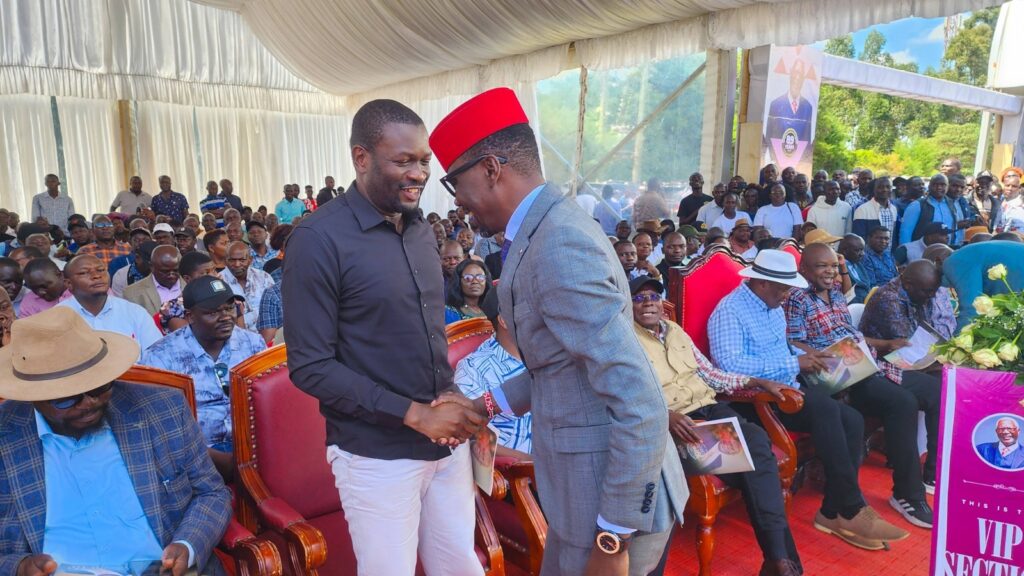 Kimilili MP Didmus Barasa and Nairobi Senator Edwin Sifuna shake hands during Mzee Patrick Wangamati’s burial ceremony as mourners and political leaders gather under a tent in Bungoma County.