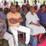 A group of mourners seated in rows at an outdoor event, with Philip Etale in a beige shirt and white trousers sitting with his arms folded at the front.