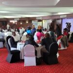 A conference room filled with journalists and health sector stakeholders seated around round tables, listening to speakers during an official event, with cameras set up at the back for coverage.