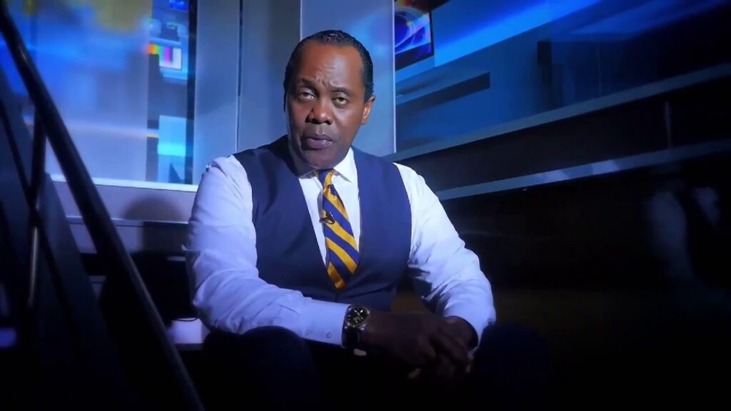 A well-dressed man in a white shirt, striped tie and navy vest sits in a dimly lit TV studio, looking directly at the camera with a serious expression.