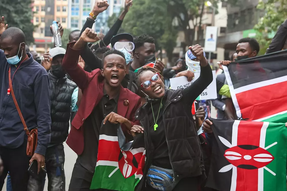 A group of young people in a street protest holding Kenyan flags, raising their fists, and shouting, with buildings visible in the background.