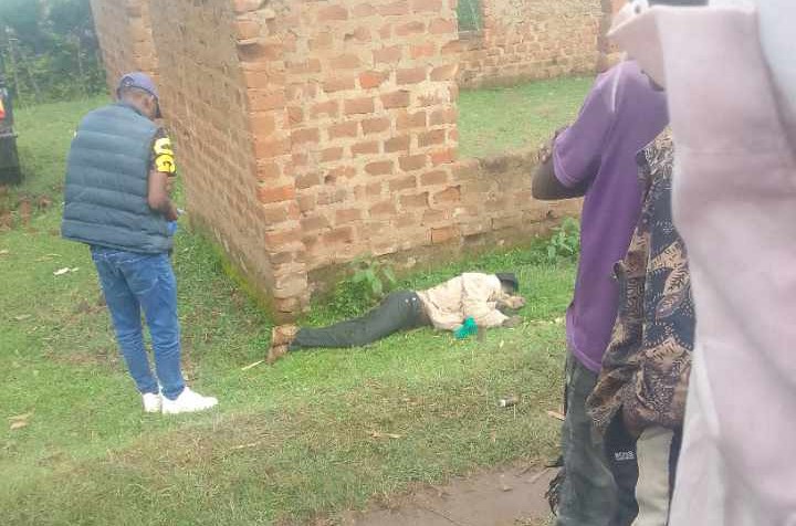 A man lies motionless on the ground beside a brick unfinished structure as a police officer and onlookers stand nearby observing the scene.
