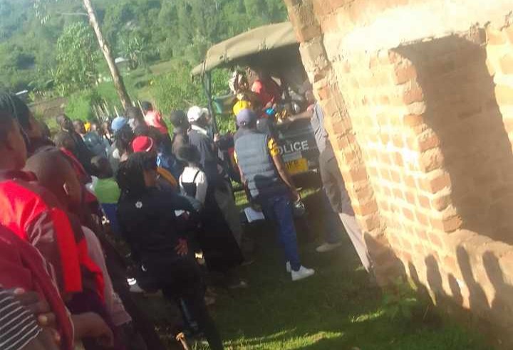 A group of police officers place a body into a police vehicle while a large crowd of people surrounds the area, watching the operation near a brick building.