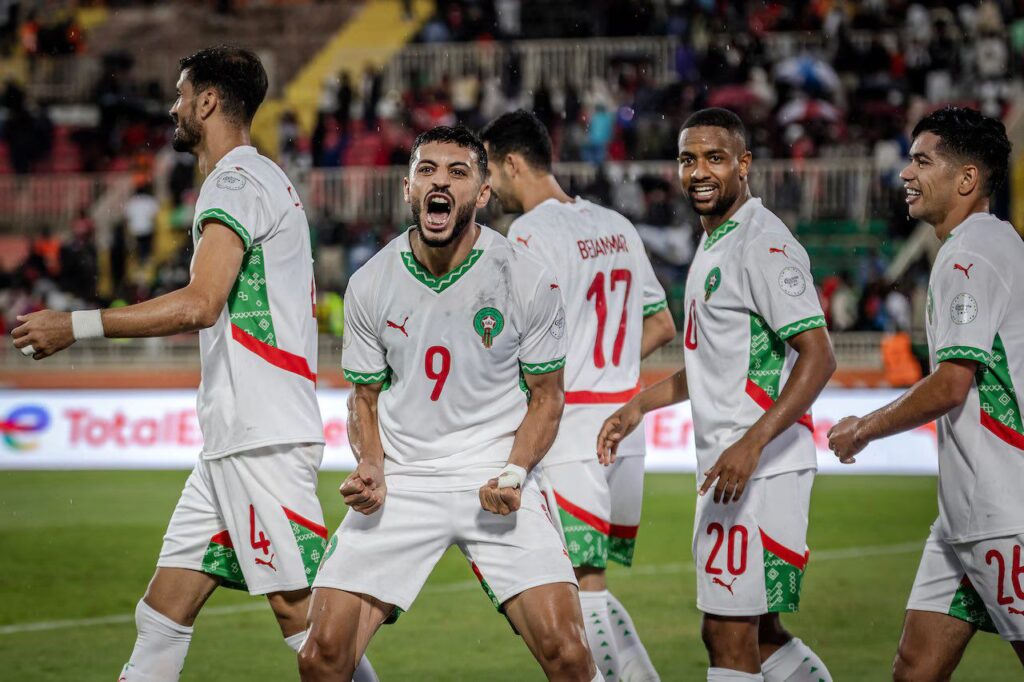Morocco national team players in white kits celebrate on the field, with one player shouting in excitement while teammates smile and gather around in a stadium setting.