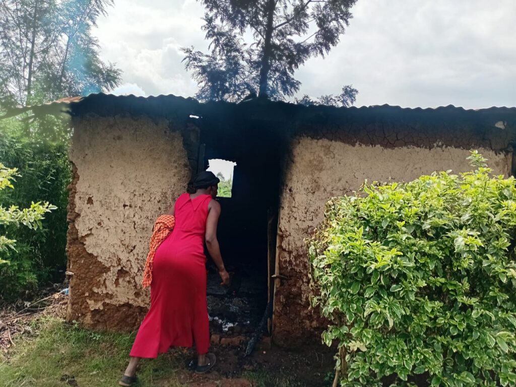 A woman in a red dress walks into a burnt mud house, examining the fire damage at the entrance.