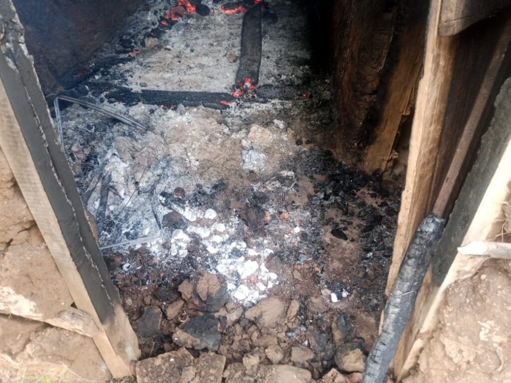 Interior of a small house showing ash-covered floor, burnt debris, and charred wooden structures after a fire.