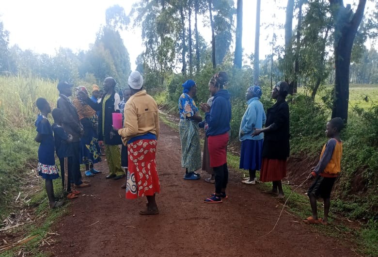 Women and children stand on a dirt road in a rural area surrounded by trees and vegetation, gathered in small groups as they react to a local incident.