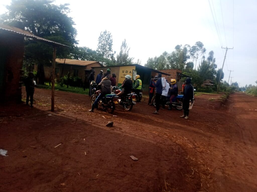A group of men, including boda boda riders with motorcycles, stand along a rural roadside near small buildings, discussing an incident in Misanga B Village.
