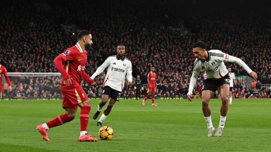 Mohamed Salah dribbling the ball during a match while two Fulham players attempt to defend against him in a crowded stadium.