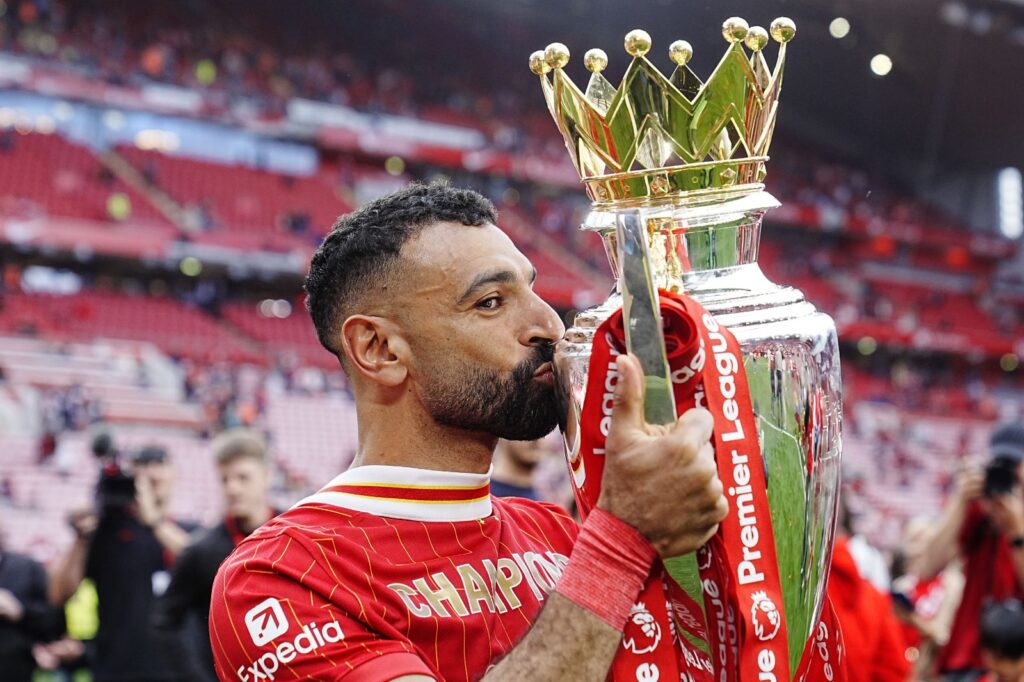 Mohamed Salah in a Liverpool jersey kissing the Premier League trophy inside a stadium after winning the title.