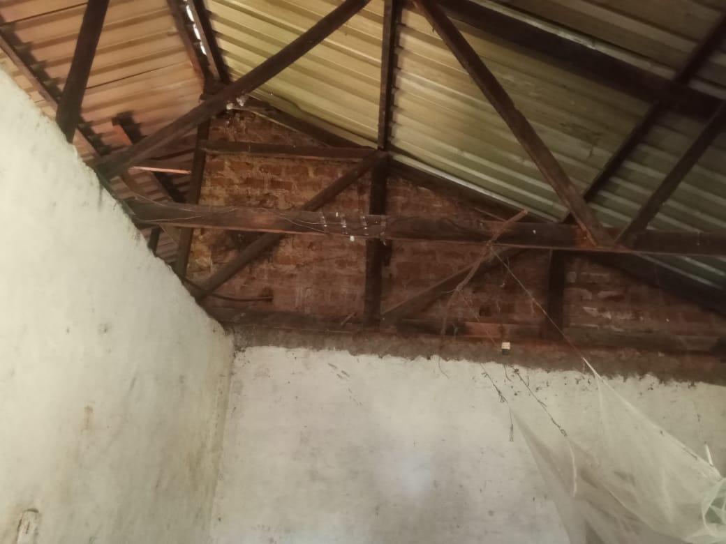 Inside of a modest room showing rusted iron roofing sheets, exposed wooden beams, and cobwebs, indicating an aging structure.