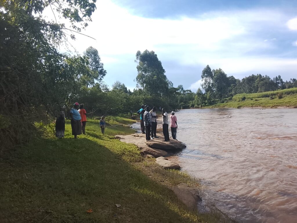 A group of men standing under trees near a muddy riverbank, discussing as they look toward River Nzoia during a search operation.
