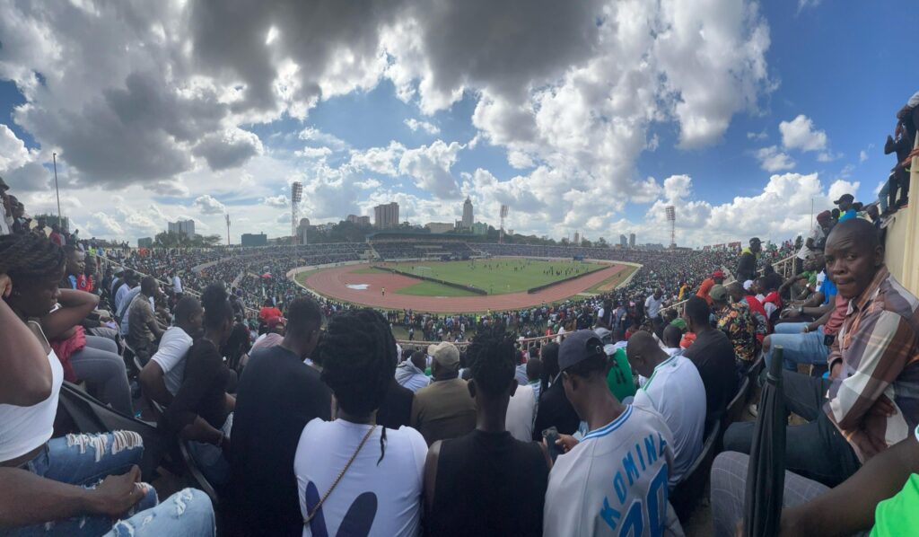 Wide view of a fully packed Nyayo National Stadium in Nairobi with thousands of fans seated around the pitch during a football match, with players visible on the field under a cloudy sky.