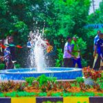 A group of students walking near a landscaped fountain with water spraying upward, surrounded by greenery and flowers on a university campus.