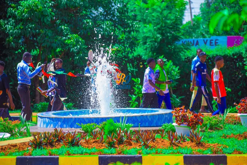 A group of students walking near a landscaped fountain with water spraying upward, surrounded by greenery and flowers on a university campus.