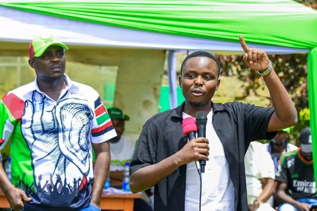 A young man speaking into a microphone and raising his hand while addressing a crowd at an outdoor political event, with another man standing beside him under a green and white tent.