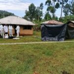 Several forensic officers in protective suits stand near a rural home in Vihiga County during an exhumation operation, with a tented area and police tape marking the scene.