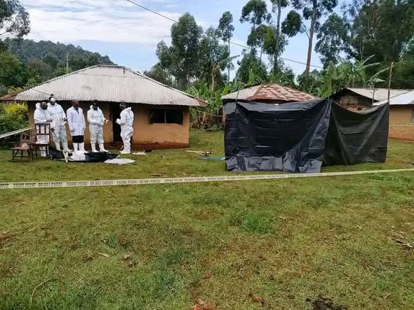 Several forensic officers in protective suits stand near a rural home in Vihiga County during an exhumation operation, with a tented area and police tape marking the scene.