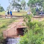 A rural roadside scene showing a concrete culvert with water flowing beneath it, surrounded by green vegetation. Several people stand nearby, including individuals on a motorcycle, as they look toward the stream during an ongoing search operation.