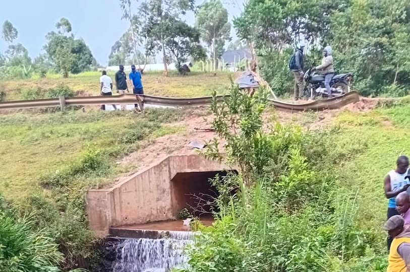 A rural roadside scene showing a concrete culvert with water flowing beneath it, surrounded by green vegetation. Several people stand nearby, including individuals on a motorcycle, as they look toward the stream during an ongoing search operation.