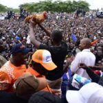 Edwin Sifuna stands amid a large, tightly packed crowd in Kisumu, holding a microphone while raising a live rooster high above his head. Supporters surround him on all sides, many cheering, taking photos, and reaching toward him in a lively political rally atmosphere.