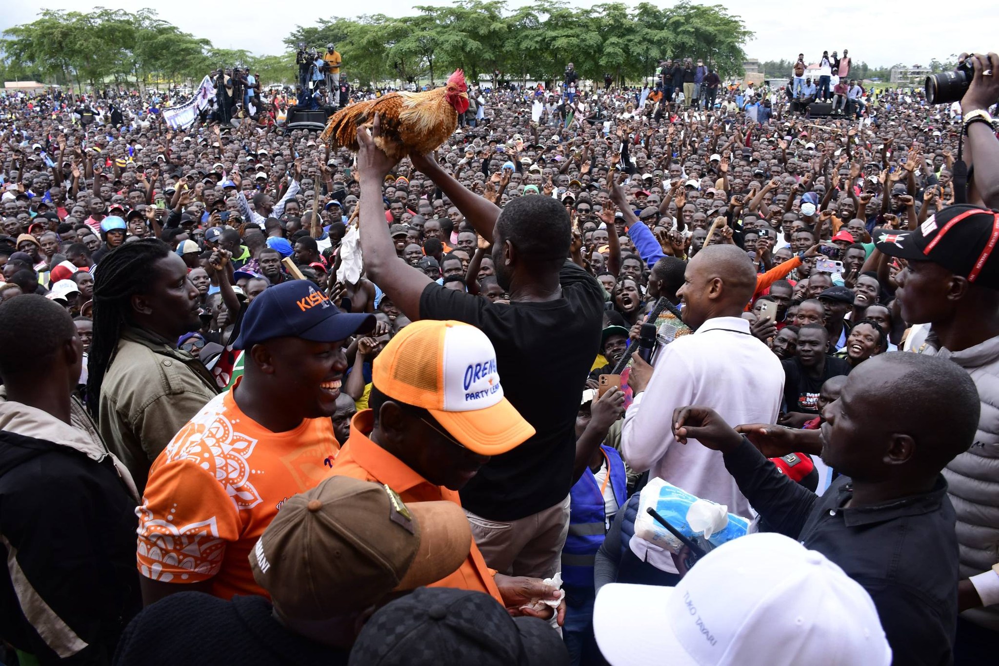 Edwin Sifuna stands amid a large, tightly packed crowd in Kisumu, holding a microphone while raising a live rooster high above his head. Supporters surround him on all sides, many cheering, taking photos, and reaching toward him in a lively political rally atmosphere.