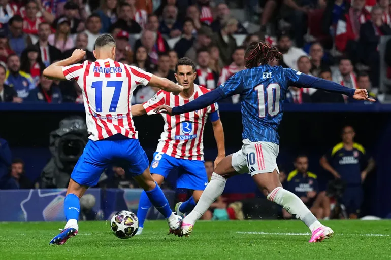 Arsenal midfielder Eberechi Eze in a blue kit challenges two Atlético Madrid defenders in red-and-white stripes while contesting the ball during a crowded Champions League match.