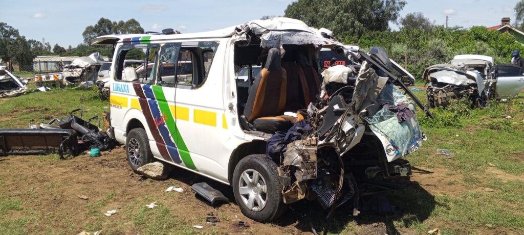 Severely damaged matatu with its front completely crushed following a road accident; debris scattered around and other wrecked vehicles visible in the background on a grassy area near the crash scene.