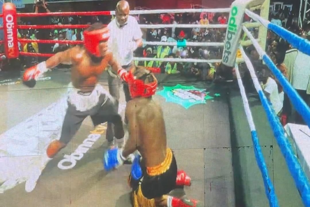 A boxing match in a crowded indoor arena shows one fighter in red headgear throwing a punch at an opponent who is down on one knee near the ropes, with a referee closely observing the action.