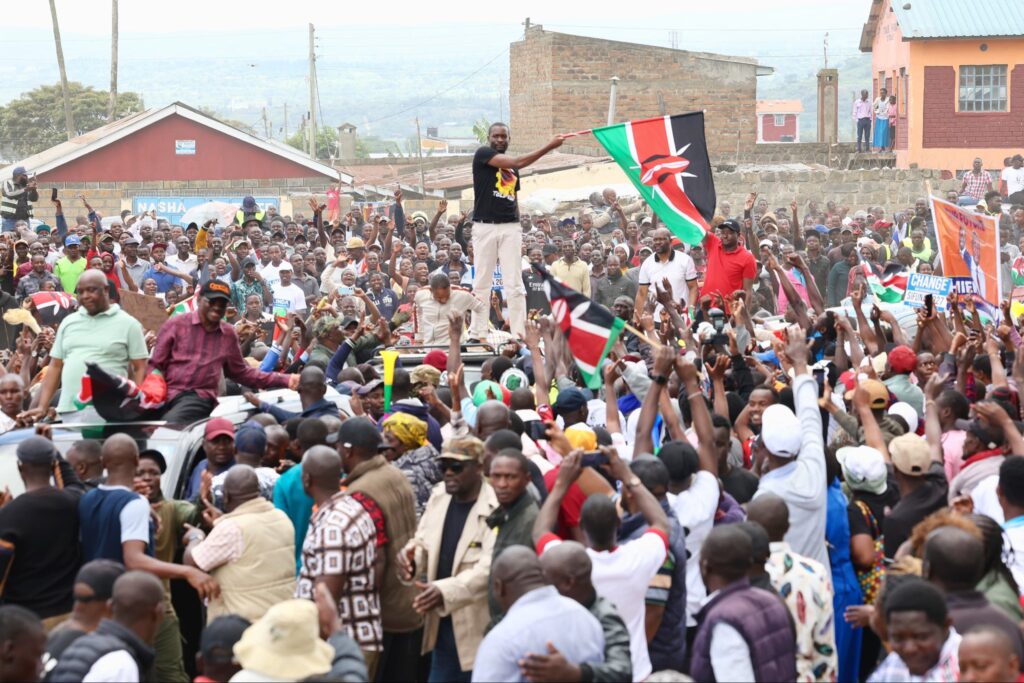 A large crowd gathers in Nakuru town as a man stands through a vehicle waving a Kenyan flag, surrounded by cheering supporters during a political rally.