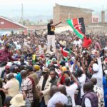 A large crowd gathers in Nakuru town as a man stands through a vehicle waving a Kenyan flag, surrounded by cheering supporters during a political rally.
