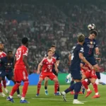A crowded penalty area scene during a Champions League match, with PSG and Bayern Munich players in blue and red kits jumping and positioning to challenge for a high ball in front of goal, as a packed stadium watches in the background.