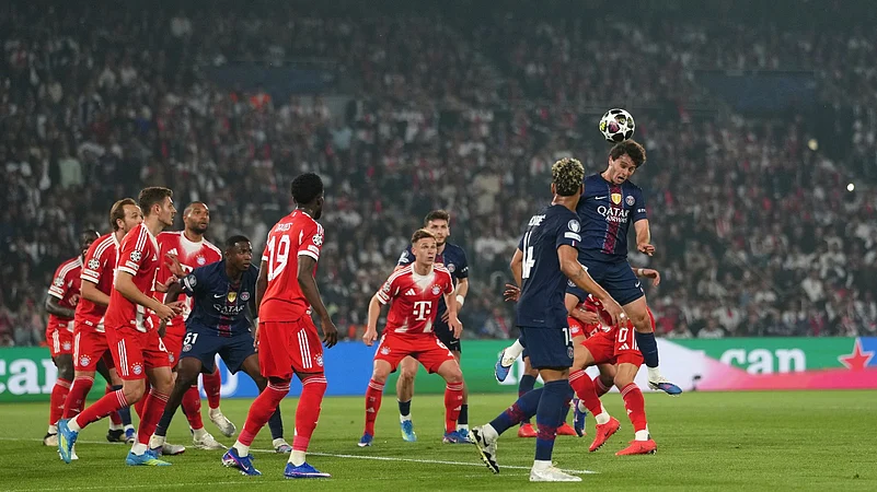 A crowded penalty area scene during a Champions League match, with PSG and Bayern Munich players in blue and red kits jumping and positioning to challenge for a high ball in front of goal, as a packed stadium watches in the background.