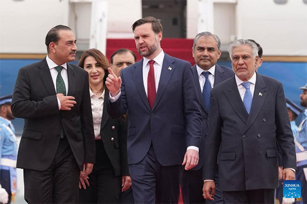 U.S. Vice President JD Vance, dressed in a navy suit and red tie, walks and gestures while speaking with a group of Pakistani officials in formal attire outside a government building during an official visit to Pakistan.