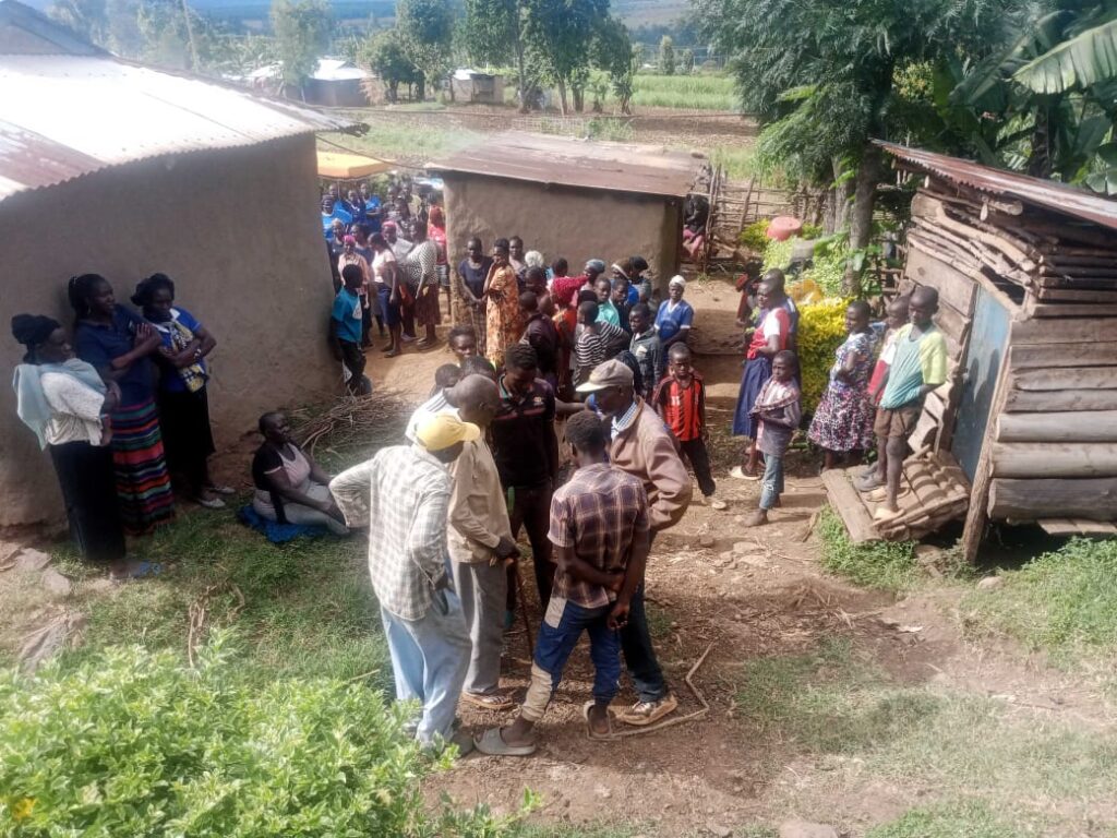 A large group of men, women, and children gather in a homestead, with some standing and others seated, reacting to an incident that has drawn community attention.
