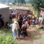 A large group of men, women, and children gather in a homestead, with some standing and others seated, reacting to an incident that has drawn community attention.