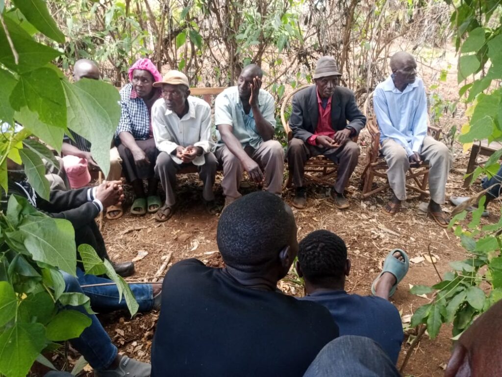 A group of men and one woman seated outdoors in a rural setting, engaged in a community meeting under trees, with others seated in front listening.