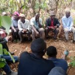 A group of men and one woman seated outdoors in a rural setting, engaged in a community meeting under trees, with others seated in front listening.