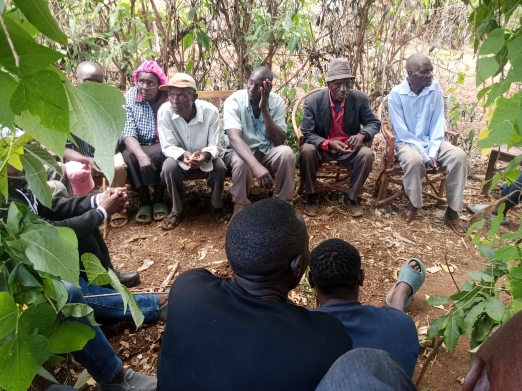 A group of men and one woman seated outdoors in a rural setting, engaged in a community meeting under trees, with others seated in front listening.