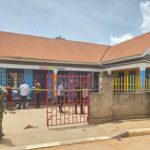 Armed security officers stand outside a gated daycare centre in Kampala, cordoned off with police tape, as investigators and bystanders gather inside following a deadly attack.