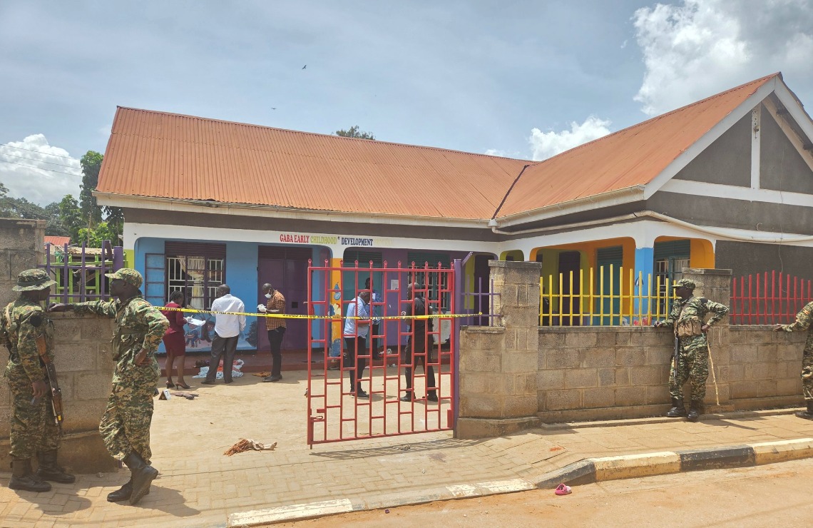 Armed security officers stand outside a gated daycare centre in Kampala, cordoned off with police tape, as investigators and bystanders gather inside following a deadly attack.