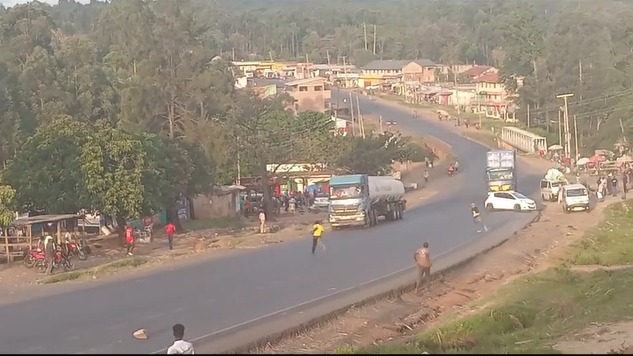 A roadside accident scene showing a tanker truck and another lorry stopped on a curved highway, with people gathered nearby and some moving toward the vehicles in a semi-rural area.