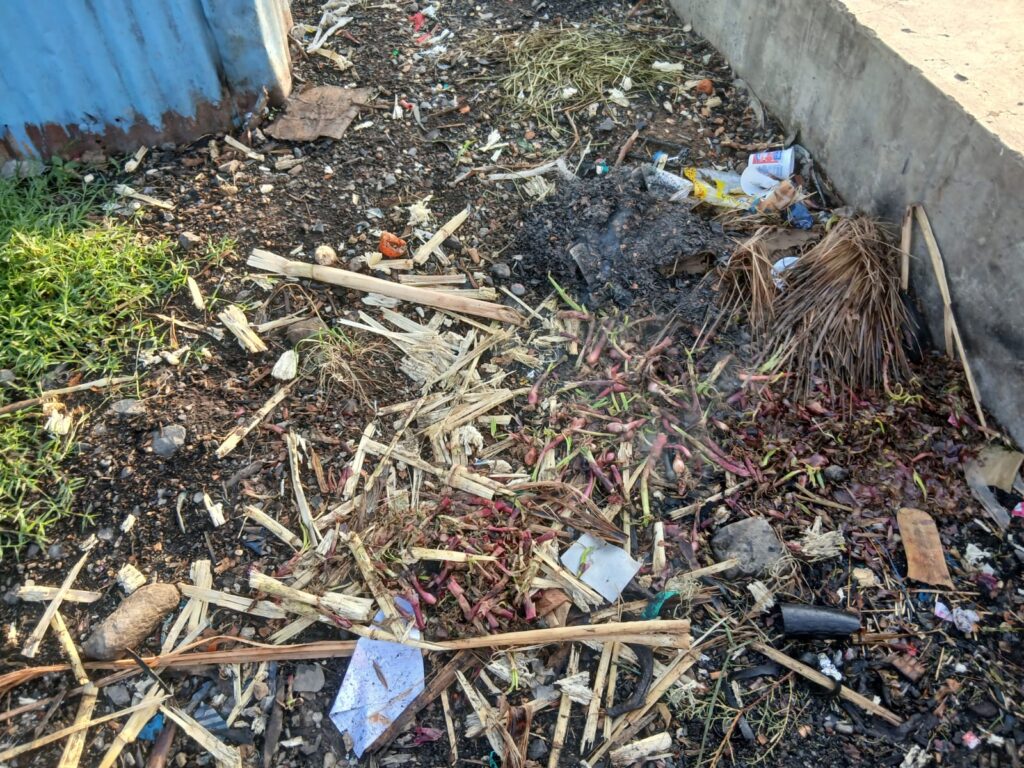 Pile of mixed garbage including organic waste, wood pieces, and plastic scattered on the ground beside a structure at Chimoi Market.