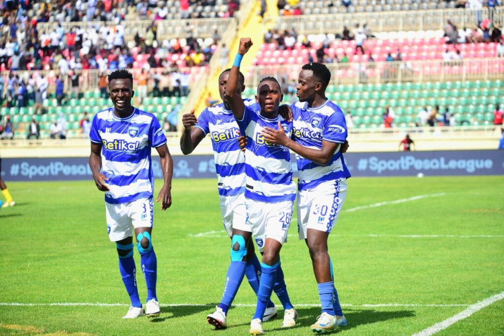 AFC Leopards players in blue and white kits celebrate on the pitch, with one player raising his fist as teammates gather around him, while fans watch from the stands in the background.