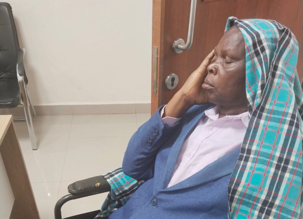 An elderly man seated in a wheelchair inside a hospital room, wearing a blue blazer and covered with a checked blanket, resting his head on his hand.