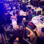 A crowded banquet hall with formally dressed attendees crouching behind chairs and tables, some on the floor, as others look around in alarm during a sudden security scare.