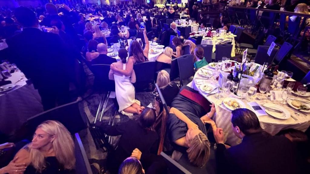 A crowded banquet hall with formally dressed attendees crouching behind chairs and tables, some on the floor, as others look around in alarm during a sudden security scare.