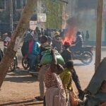 A street scene with a group of people surrounding a burning motorcycle. Thick smoke rises into the air as onlookers, some carrying bags, watch from nearby. Buildings and shops line the background, suggesting an urban setting.