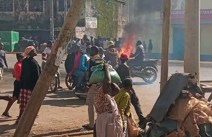 A street scene with a group of people surrounding a burning motorcycle. Thick smoke rises into the air as onlookers, some carrying bags, watch from nearby. Buildings and shops line the background, suggesting an urban setting.